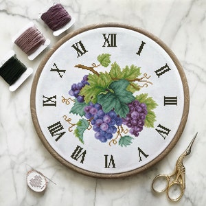 Puede incluir: Un aro de bordado blanco con una esfera de reloj y un diseño de vid. La esfera del reloj tiene números romanos y la vid está en tonos de morado y verde.