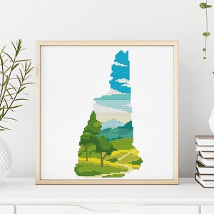May include: A cross-stitch pattern of a landscape scene in the shape of the state of New Hampshire. The scene features a blue sky with white clouds, green trees, a dirt road, and a field of grass.