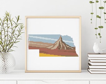 May include: A framed cross-stitch design of the state of Nebraska with a mountain range in the background. The design is in shades of brown, red, yellow, and blue.
