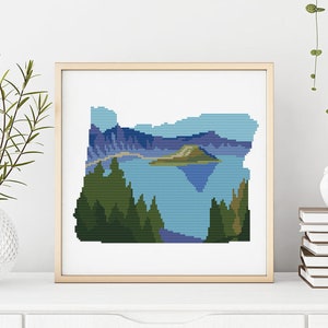 May include: A cross-stitch pattern of the state of Oregon, featuring a blue lake with an island in the center, surrounded by green trees and mountains.