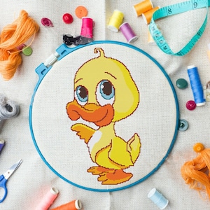 Puede incluir: Un patito amarillo con grandes ojos azules y un pico naranja está bordado en un lienzo blanco en un aro de bordado azul.