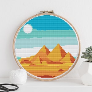 May include: A cross-stitch embroidery hoop with a colorful depiction of the pyramids in a desert landscape. The sky is blue with white clouds and a white sun. The pyramids are orange, yellow, and red.