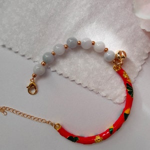 May include: A bracelet with a red, green, and yellow enamel band and a string of light blue jade beads. The bracelet has a gold clasp and a small gold bell charm.