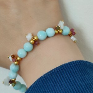 May include: A delicate bracelet featuring light blue spherical beads interspersed with gold-colored beads and small, colorful gemstones. The bracelet also includes small white flower-shaped charms. The bracelet is designed to be worn on the wrist.