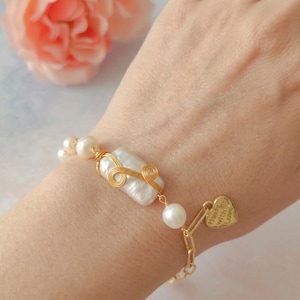 May include: A bracelet featuring white pearls and a central rectangular baroque pearl wrapped in gold-colored wire. The bracelet includes a gold-colored chain segment and a small heart-shaped charm engraved with the text Hand with Love.