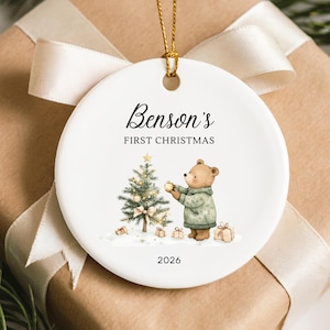 May include: A round white ceramic ornament featuring a gold string, printed with the text Benson's FIRST CHRISTMAS and 2026. The design includes a brown bear wearing a green sweater decorating a Christmas tree with small gifts on the snow.