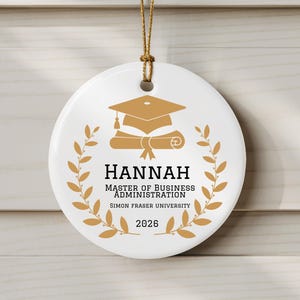 May include: White ceramic graduation ornament with gold-colored graphics and text. The ornament features a graduation cap, diploma, and laurel wreath design. Text includes the name "HANNAH", "MASTER OF BUSINESS ADMINISTRATION", "SIMON FRASER UNIVERSITY", and the year "2026".