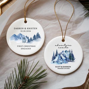 Peut inclure: Deux ornements ronds en céramique blanche avec des cordons dorés. L'un présente des montagnes à l'aquarelle bleue, le texte DARREN & KRISTEN, 10.28.2026, et FIRST CHRISTMAS ENGAGED. L'autre montre des montagnes bleues, adventure awaits, SCOTT & SHANNON, 11-15-2026.