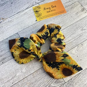 Könnte beinhalten: Ein Haargummi mit Sonnenblumenmuster und Schleife. Das Design zeigt gelbe Sonnenblumen auf schwarzem Hintergrund mit weißen Punkten. Das Haaraccessoire hat eine Schleife. Eine Visitenkarte im Hintergrund lautet "Honey Bea Hair & Co Quality & Cute Scrunchies & Hair Accessories."