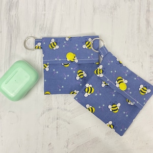 Könnte beinhalten: Zwei blaue Stoffbeutel mit einem Bienenmuster und einer gelben Mitte. Die Beutel haben einen silbernen Ring oben angebracht und sind zugeklappt.