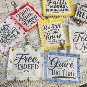 May include: A collection of square fabric keychains with scalloped edges, each featuring black printed text with religious phrases like Faith Runs Deep, Be Still and Know, Free Indeed, and Grace Did That, attached to silver metal swivel clasps.