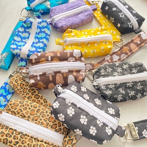 Op de afbeelding: Een assortiment kleine ritszakjes in verschillende patronen en kleuren, waaronder bruin met paddenstoelen, zwart met witte pootjes, zwart met witte sterren en luipaardprint. De zakjes zijn gemaakt van stof en hebben een ritssluiting.