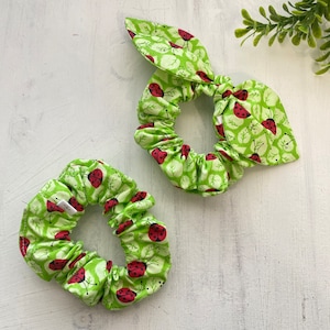 May include: Two green and red scrunchies with a ladybug print. The scrunchies are made of fabric and have a gathered look.