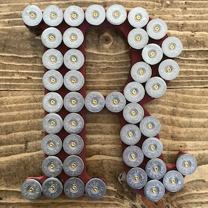 Könnte beinhalten: Ein roter Holzbuchstabe R, der mit silbernen Schrotpatronenhülsen verziert ist. Die Hülsen sind in einem kreisförmigen Muster um den Buchstaben angeordnet, wobei auf einigen Hülsen die Worte "12 GA FEDERAL" zu sehen sind.