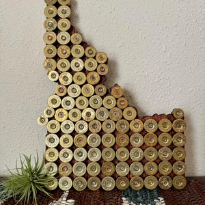 May include: A wooden wall art piece in the shape of Idaho, decorated with numerous brass bullet casings. The casings are arranged closely together, creating a textured, metallic surface. The artwork is mounted on a wooden base, showcasing a rustic aesthetic.