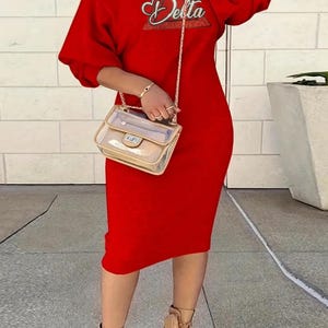 Könnte beinhalten: Ein rotes Midikleid mit langen Ärmeln und dem Aufdruck "Delta" auf der Vorderseite. Das Kleid wird mit beigefarbenen Stiefeln und einer transparenten Handtasche mit goldener Kette kombiniert.