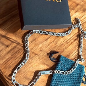 May include: A silver-toned metal chain necklace featuring a rectangular bar pendant engraved with the name Vanessa. The necklace is displayed alongside a dark blue fabric drawstring pouch and a black box with the gold text DVINO printed on top.