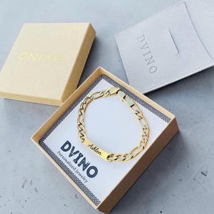 May include: A gold-toned Figaro chain bracelet featuring a rectangular identification plate engraved with the name Ashlen. The bracelet is presented in a square cardboard box labeled DVINO Personalized Jewelry.