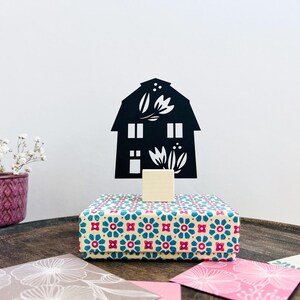 Könnte beinhalten: Eine schwarze, hausförmige Dekoration mit floralen Ausschnitten steht auf einem kleinen Holzblock auf einer gemusterten Box. Die Box hat einen cremefarbenen Hintergrund mit blauen und roten floralen Designs. Ein violetter Topf mit weißen Blumen und dekorativen Karten sind ebenfalls im Bild.