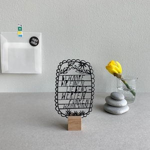 Könnte beinhalten: Schwarzes Metallschild mit dem deutschen Satz „BEWAHRE DIR EINEN LIEBENS-WERTEN FUNKEN WAHNSINN“ auf einem Holzsockel. Eine gelbe Blume im Glas und ein Steinhaufen sind ebenfalls zu sehen.