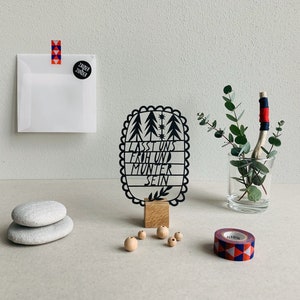 Könnte beinhalten: Schwarz-weißes Papier-Ausschnitt einer Waldszene mit dem Text "Lasst uns froh und munter sein" in einem schwarzen ovalen Rahmen. Der Ausschnitt steht auf einem Holzsockel und es gibt Holzperlen und eine Rolle Washi-Tape auf einer weißen Oberfläche.