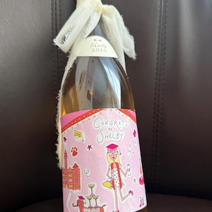 May include: A glass wine bottle containing a light-coloured liquid, finished with a cream screw cap and a frayed white ribbon. The label displays a pink background with illustrations of a graduate, a dachshund, and the text Congrats to Shelby.