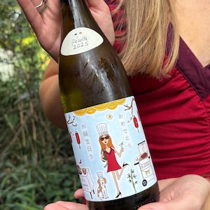 Puede incluir: Una botella de vino con tapón de lámina dorada y una etiqueta blanca con una ilustración dibujada a mano de una persona con vestido rojo, un perro y una parrilla. La etiqueta incluye el texto "Récolte 2025" y caracteres japoneses.