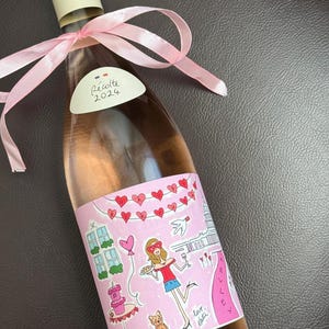 May include: A bottle of rosé wine with a pink label featuring a cartoon illustration of a woman, hearts, and the text "Kelley". The bottle is tied with a pink ribbon and has a white label with "Récolte 2024".
