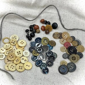 May include: A collection of assorted buttons in various sizes and colors, including cream, brown, black, and blue. The buttons are made of different materials and arranged on a light-colored fabric with a gray thread.