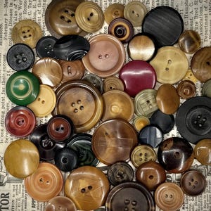 May include: A collection of assorted round buttons in various sizes and colors, including shades of brown, black, green, and burgundy. The buttons feature different designs, some with two or four holes, and are scattered across a printed paper surface.