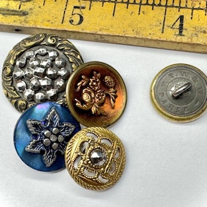 May include: A collection of vintage buttons in various designs and colors. The buttons include ornate metalwork, floral patterns, and a blue starburst design. A wooden ruler is in the background.