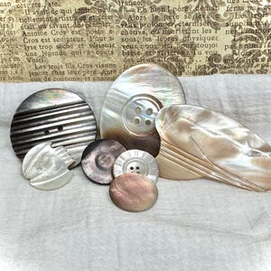 May include: A collection of various iridescent mother-of-pearl buttons in different shapes and sizes. The buttons include round, oval, and irregular shapes, with varying textures and buttonhole designs. The buttons are displayed on a white fabric background.