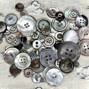 May include: A collection of assorted round buttons in various shades of gray, charcoal, and iridescent silver. The buttons feature different sizes, ranging from small to large, with two or four holes, and some include a central recessed design.