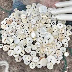 May include: A large collection of assorted white and off-white buttons in various sizes and shapes, including two-hole and four-hole designs. The buttons feature smooth, iridescent, and matte finishes, scattered together in a dense, overlapping pile.