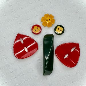 May include: A collection of colorful buttons and a green bar-shaped object. The buttons include a yellow flower-shaped button, two red buttons with gold centers, and a dark green button with a gold center. Two red, triangular buttons are also present.