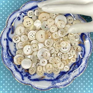 May include: A collection of assorted round white and cream-colored buttons made of mother-of-pearl, featuring various sizes and hole configurations, contained within a scalloped porcelain dish with blue and gold floral patterns.
