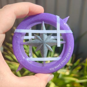 Może przedstawiać: Okrągły, fioletowy, błyszczący kompas z żywicy z srebrną gwiazdą w środku. Kompas ma białe gumki przechodzące przez niego i napis "YOU MAKE STRAY KIDS STAY" wokół krawędzi.