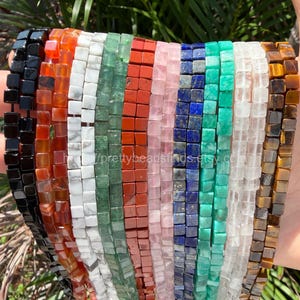 May include: A hand displays multiple strands of square gemstone beads in various colors. The beads include black, orange, white, green, red, pink, blue, and clear. Each bead is approximately 0.15 inches square. The beads are strung together, creating a colorful display.