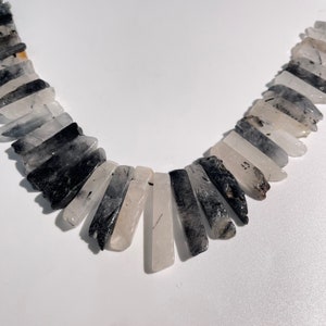 Könnte beinhalten: Eine Halskette aus schwarzen und weißen Kristallperlen. Die Perlen sind lang und schmal und in Fächerform angeordnet. Die Halskette ist ein Statement-Stück, das perfekt für einen besonderen Anlass wäre.