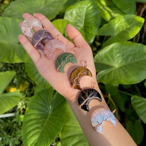 May include: Six crescent moon pendants with copper wire tree designs. The pendants are made of various stones, including amethyst, green aventurine, tiger's eye, black obsidian, and opal.