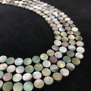 Op de afbeelding: Close-up van een ketting gemaakt van iriserende, platte, ronde kralen. De kralen hebben een parelmoerachtige uitstraling met hints van groen, paars en zilver. De ketting is gerangschikt in meerdere strengen tegen een donkere achtergrond.
