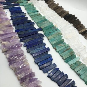 May include: A collection of gemstone beads in various colours, including amethyst, lapis lazuli, green aventurine, clear quartz, and smoky quartz. The beads are all cut into long, thin, rectangular shapes with pointed ends, resembling small crystals.