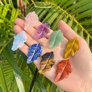 May include: Assortment of carved gemstone leaf pendants in various colors, including pink, purple, green, blue, yellow, brown, and red. Each leaf pendant is intricately detailed and displayed against a natural green background.