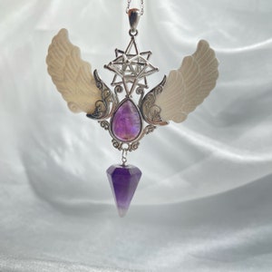 May include: A silver pendant with a purple amethyst crystal, a geometric star design, and two white wings. The pendant is on a silver chain.