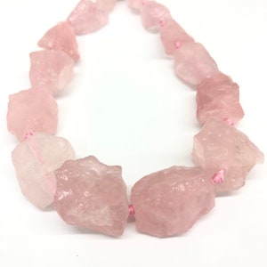 Könnte beinhalten: Eine Kette aus rosafarbenen Rosenquarzperlen, jede Perle hat eine raue, unregelmäßige Form und ist auf einem rosafarbenen Faden aufgefädelt.
