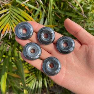 May include: Five black hematite donut shaped stones with a polished finish. The stones are arranged in a hand.