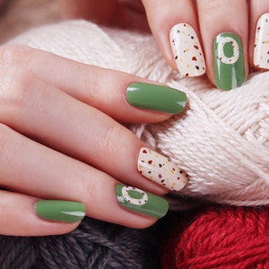 May include: Close-up of hands with green and beige nail polish. The nails are decorated with a white circle and small brown and beige dots. The hands are holding a ball of white yarn.
