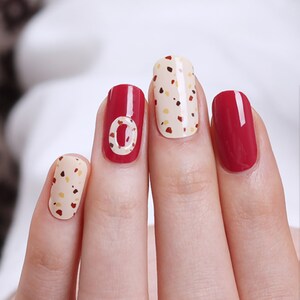 May include: Close-up of a hand with five fingernails painted with different nail polish colors and designs. The middle fingernail is painted red with a white circle design. The ring fingernail is painted red. The index fingernail is painted with a beige base and brown and white polka dots. The thumb and pinky fingernails are painted with the same beige base and brown and white polka dots.