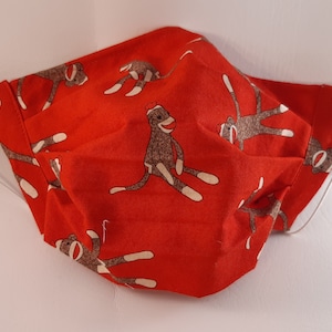May include: Red fabric face mask with a pattern of brown sock monkeys. The mask has white elastic ear loops.