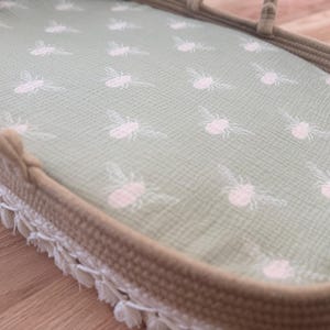 May include: A light green baby bassinet with a white bee pattern on the fabric. The bassinet has a woven brown border and a white fringe trim. The bassinet is on a wooden surface.